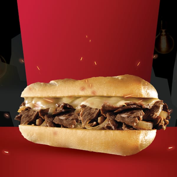 A Texas Burger Drive-In cheesesteak sandwich with melted cheese and grilled onions on a red and black background.