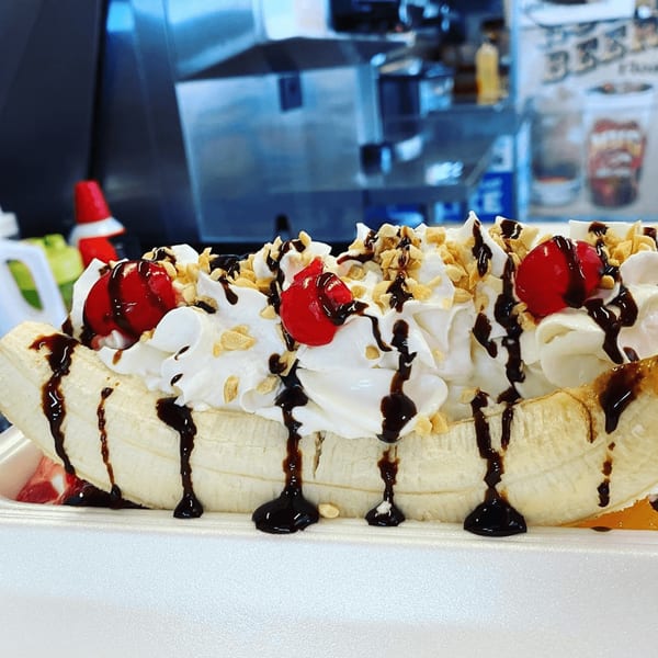 A banana split with whipped cream, chocolate syrup, cherries, and nuts in a white container.